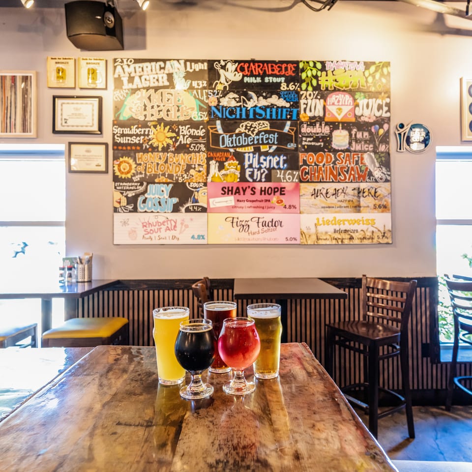 Colorful beer flights sit on a wooden restaurant table beneath a chalkboard menu displaying craft beers and cocktails.