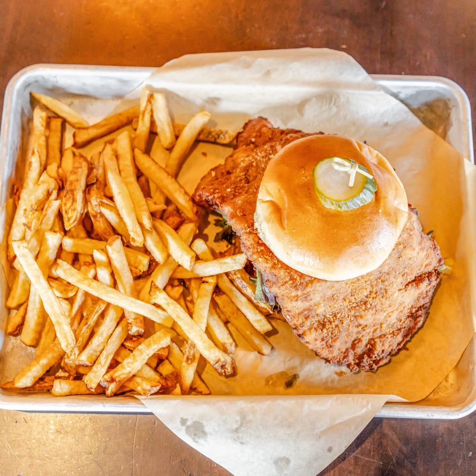 Crispy fried chicken sandwich topped with pickles beside golden French fries in a paper-lined metal tray.