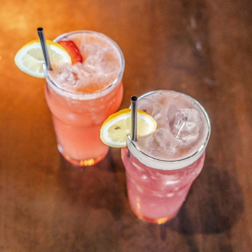 Two pink citrus cocktails with ice, lemon garnishes, and black straws on a wooden restaurant table.