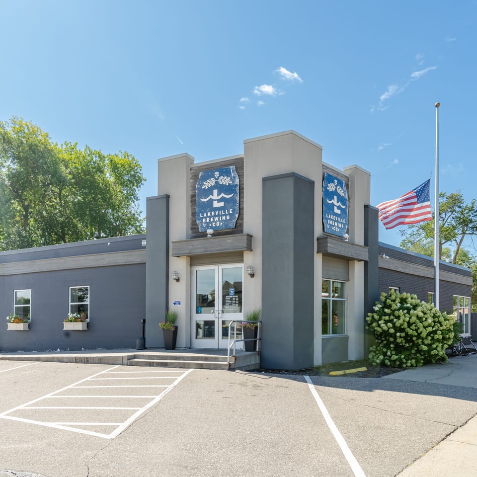 Lakeville Brewing Co. restaurant exterior with blue signage, American flag, landscaped entrance, and sunny parking lot.