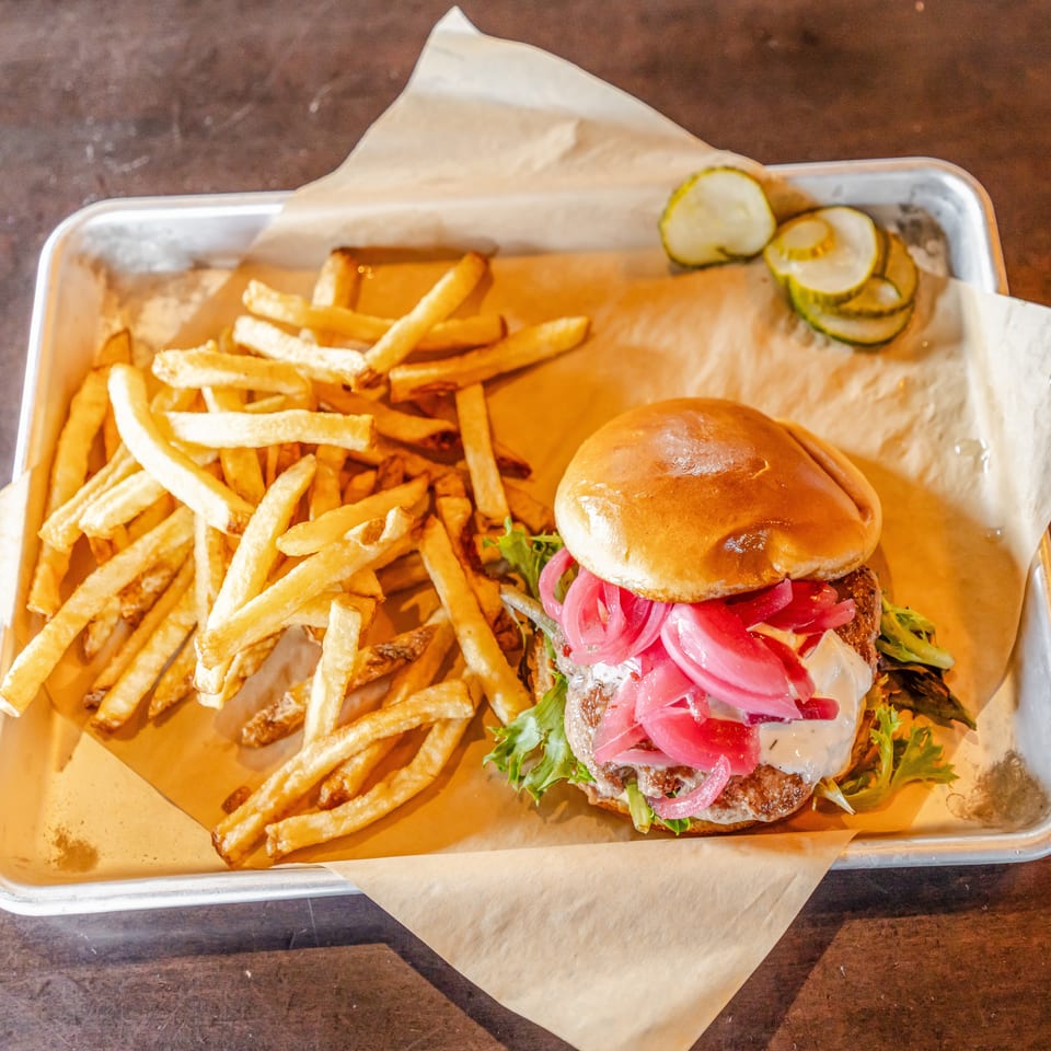 Cheeseburger topped with pickled onions and lettuce served with fries and pickle slices on a paper-lined metal tray.