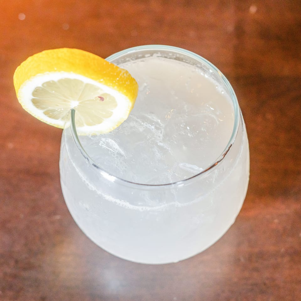 Clear cocktail with ice and a lemon slice garnish in a stemless glass on a wooden table.