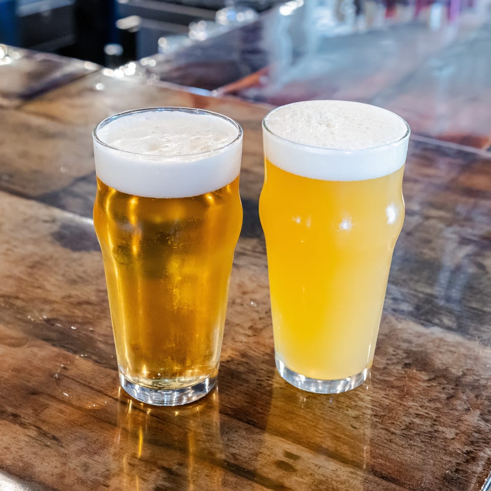 Two golden beers with frothy white heads sit on a polished wooden bar in a restaurant.