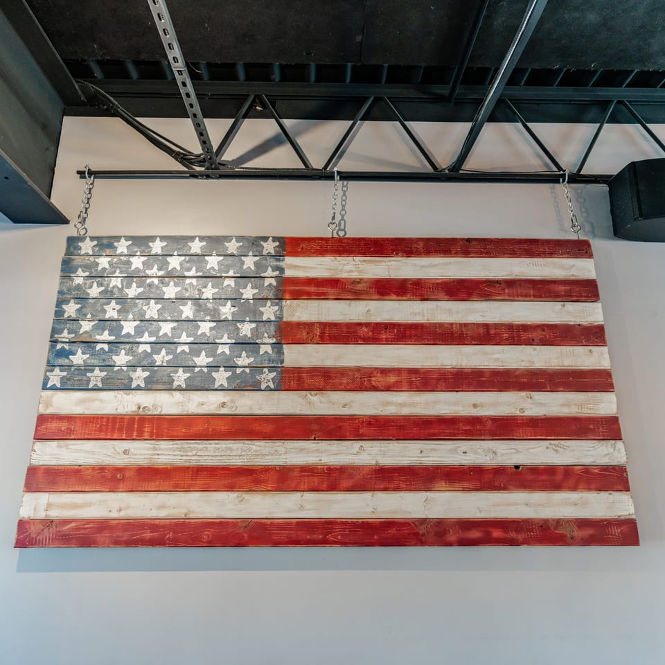 Distressed wooden American flag hangs from chains beneath an exposed industrial ceiling.