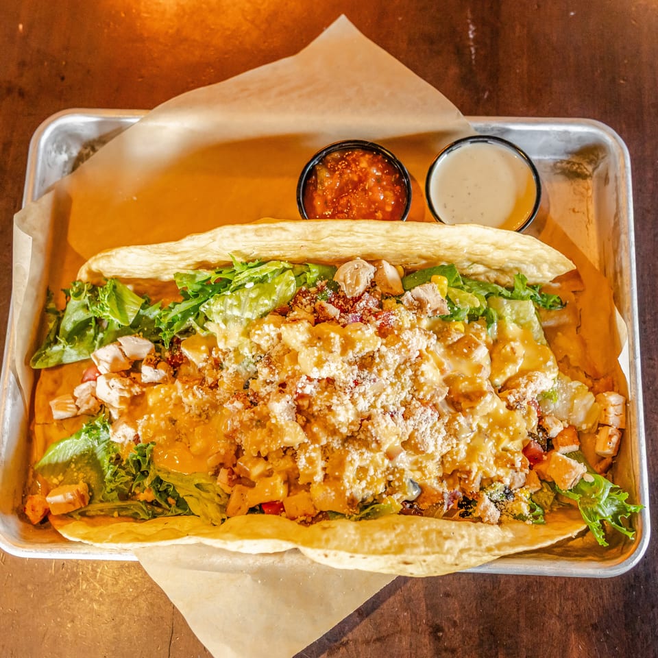 Loaded flatbread topped with grilled chicken, lettuce, vegetables, cheese, and sauces in a metal serving tray.