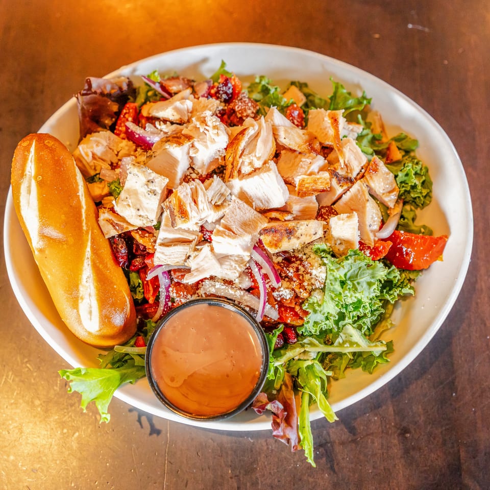 Grilled chicken salad with mixed greens, vegetables, cheese, bread roll, and creamy dressing in a white bowl.