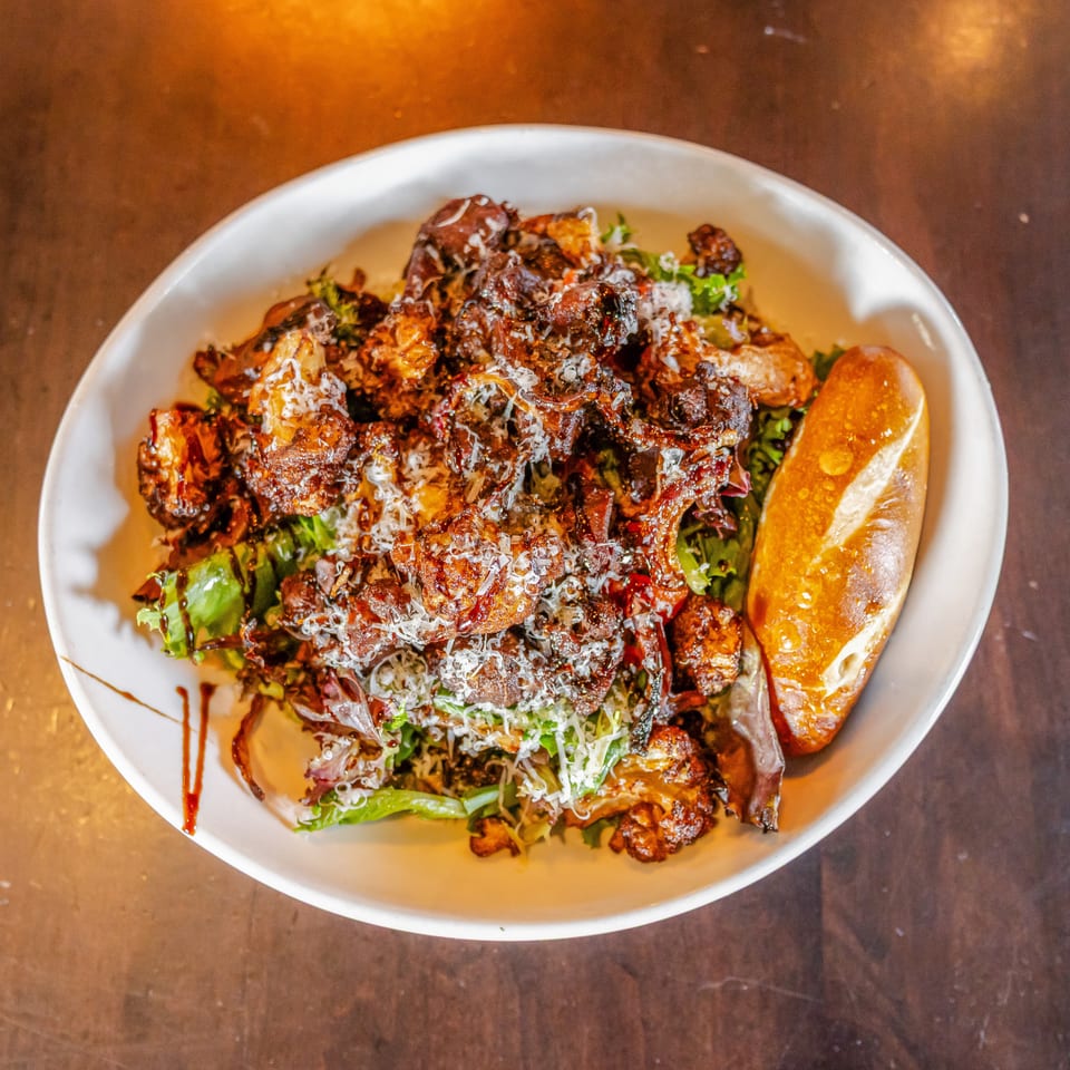 Overhead view of a white bowl filled with crispy glazed meat, greens, shredded cheese, and a lemon wedge.