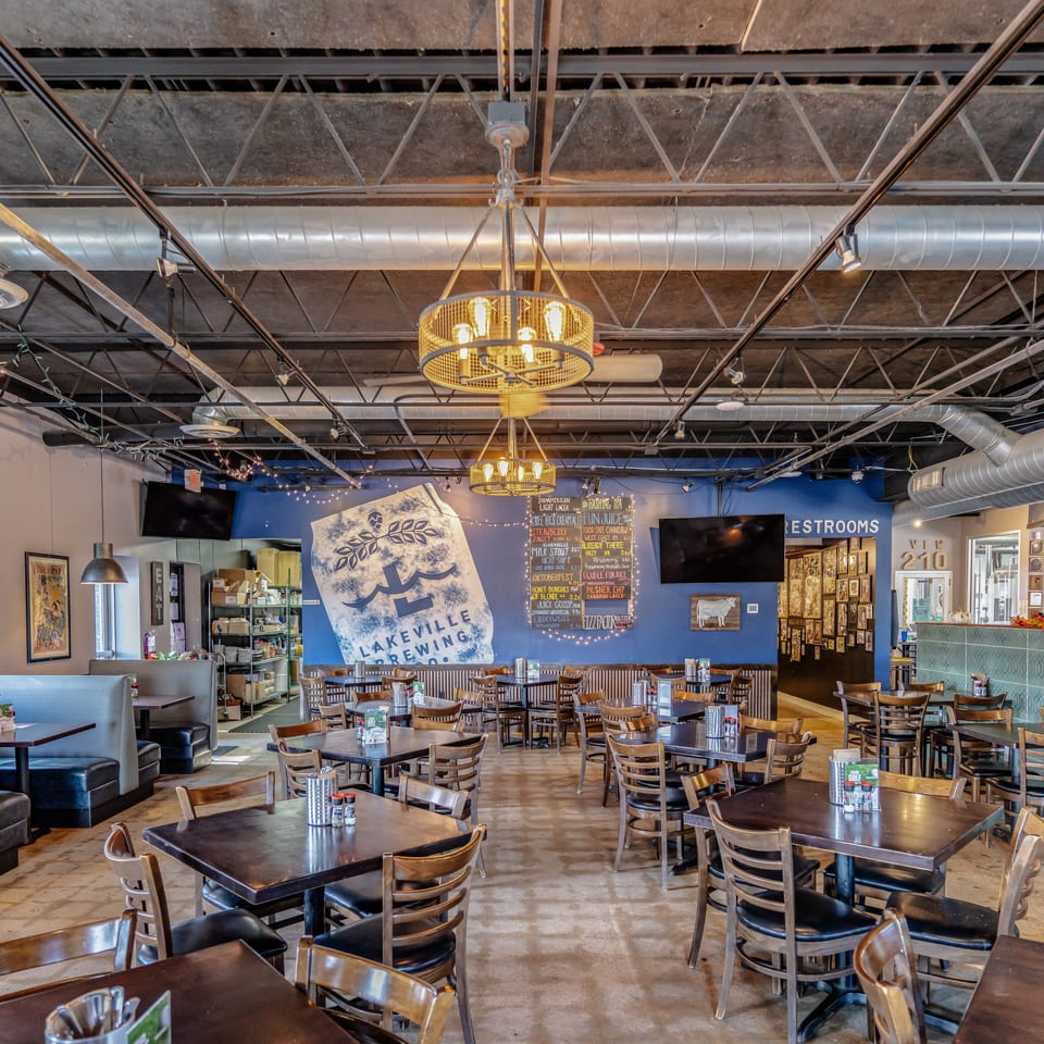 Spacious brewery restaurant dining room with wooden tables, booths, exposed ductwork, pendant chandeliers, televisions, and Lakeville Brewing Co. signage.