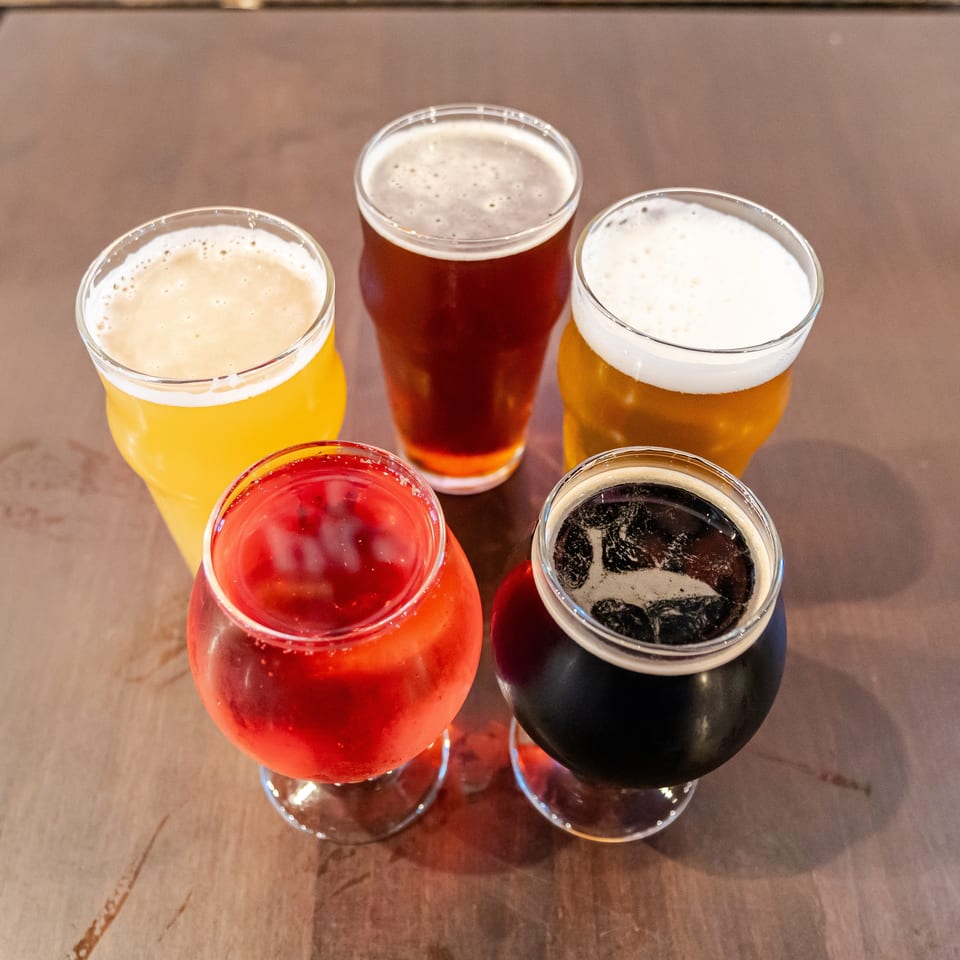Six colorful beer glasses, including pale, amber, red, and dark brews, arranged on a wooden restaurant table.