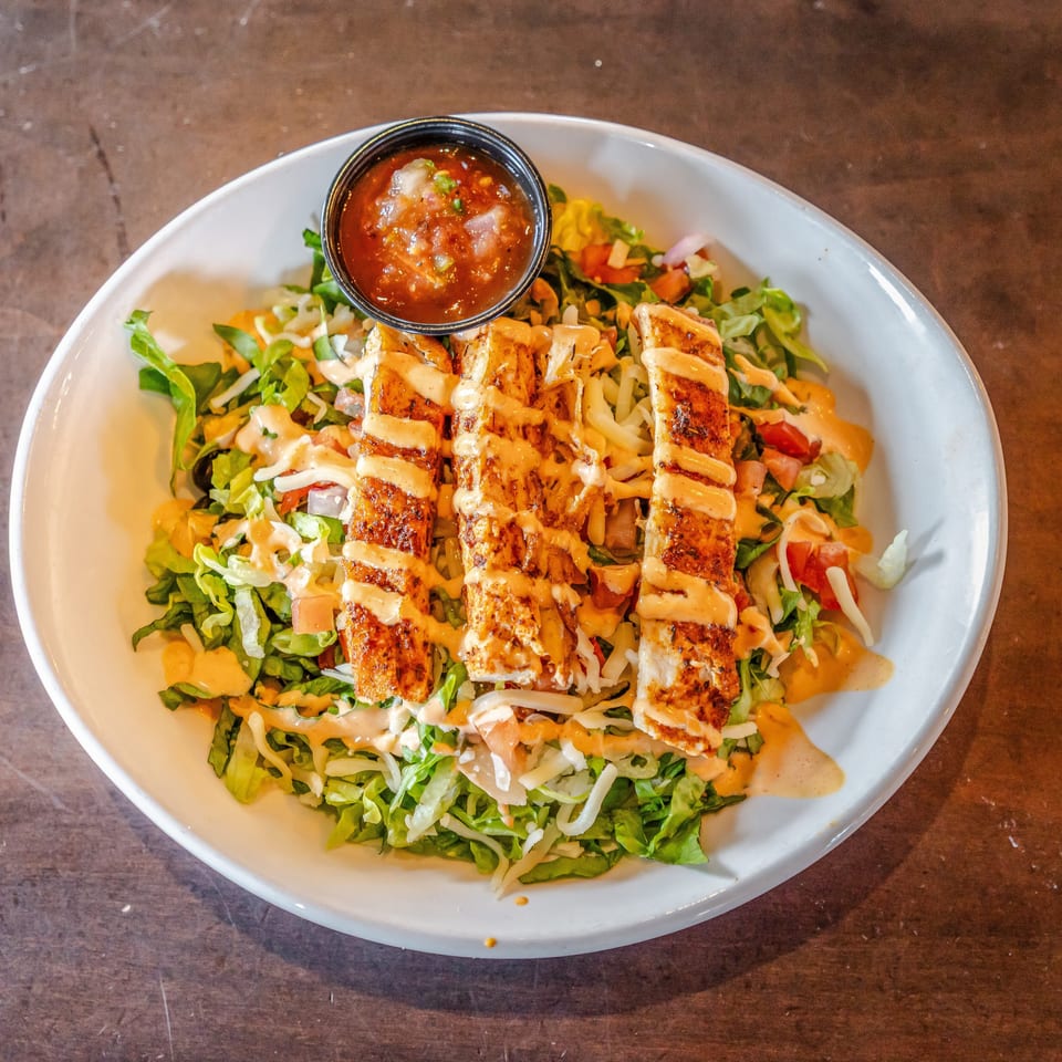 Grilled chicken strips top a chopped lettuce salad with cheese, tomatoes, creamy dressing, and a side of salsa.