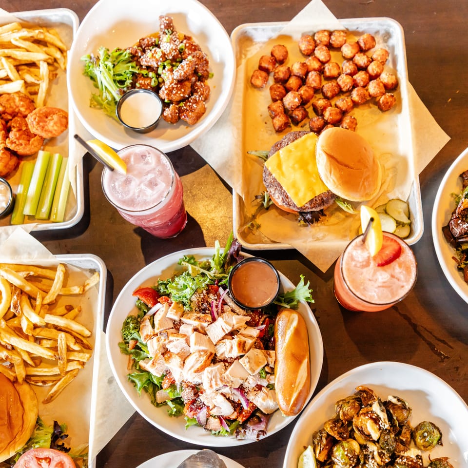 Overhead spread of burgers, fries, fried chicken, sauced bites, salads, roasted Brussels sprouts, dipping sauces, and colorful drinks on a restaurant table.