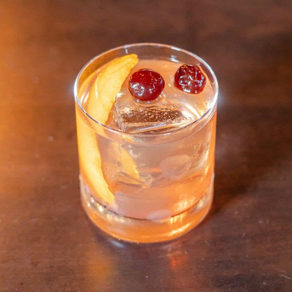 Amber cocktail with a large ice cube, orange peel, and two cherries in a rocks glass on a wooden bar.