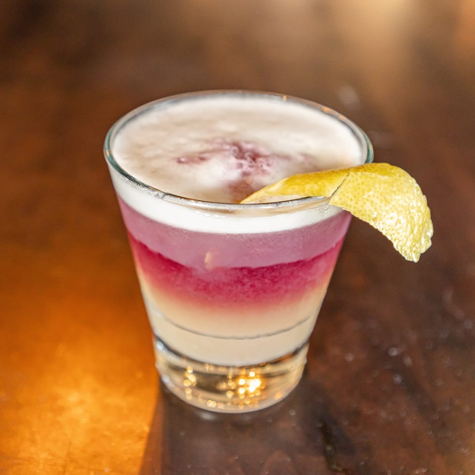 Layered pink cocktail topped with creamy foam and garnished with a curled lemon peel on a warmly lit bar.