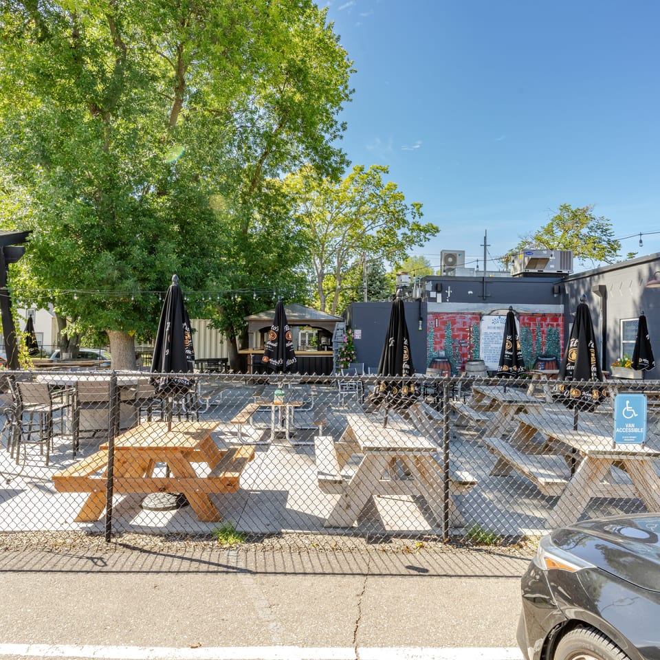 Outdoor restaurant patio with picnic tables, closed branded umbrellas, string lights, trees, and an accessible parking sign.