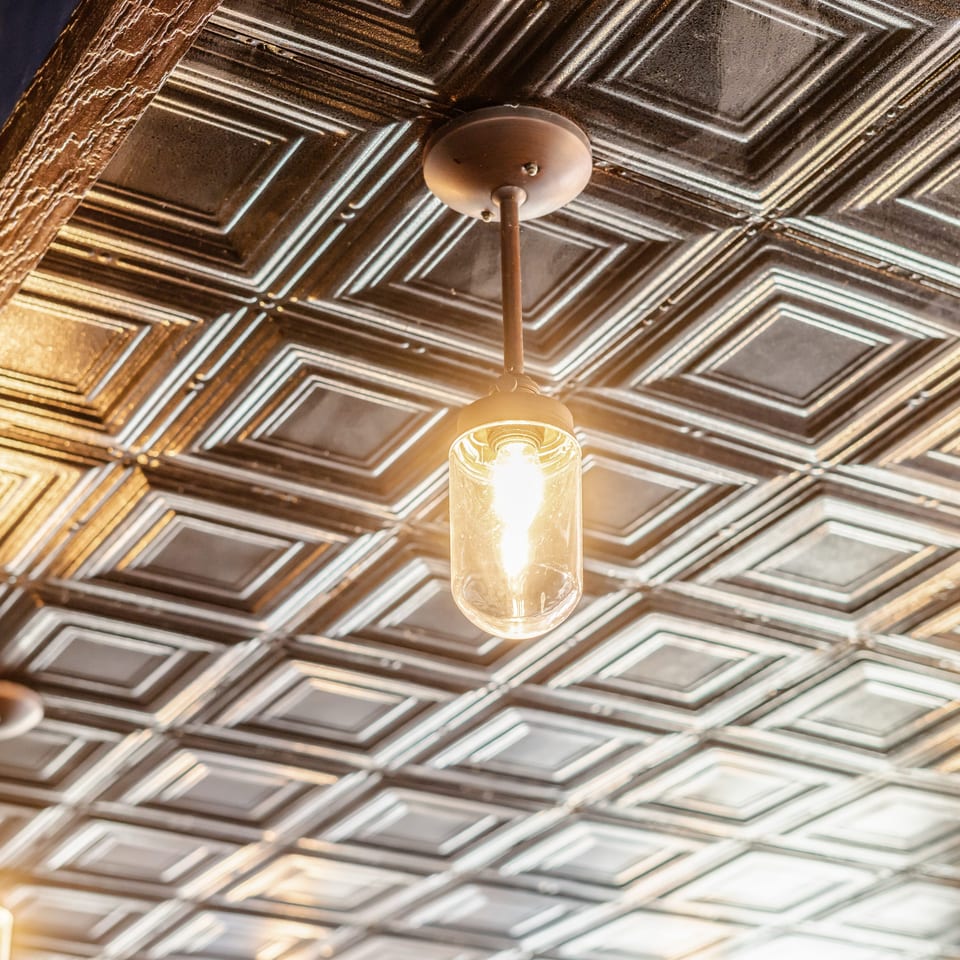 Warm pendant lights hang from an ornate, geometric pressed-tin ceiling in a restaurant interior.