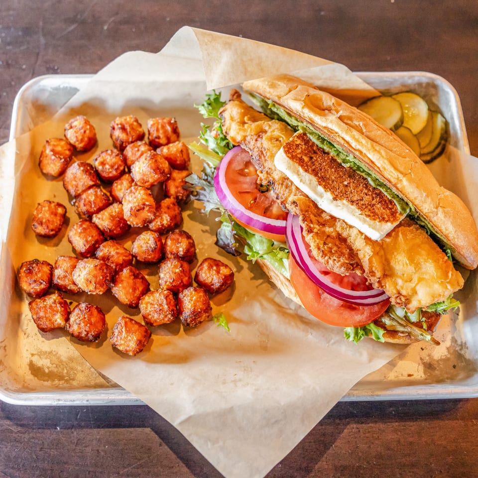 Fried chicken sandwich with lettuce, tomato, onion, and cheese served with roasted potato bites and pickles on a metal tray.