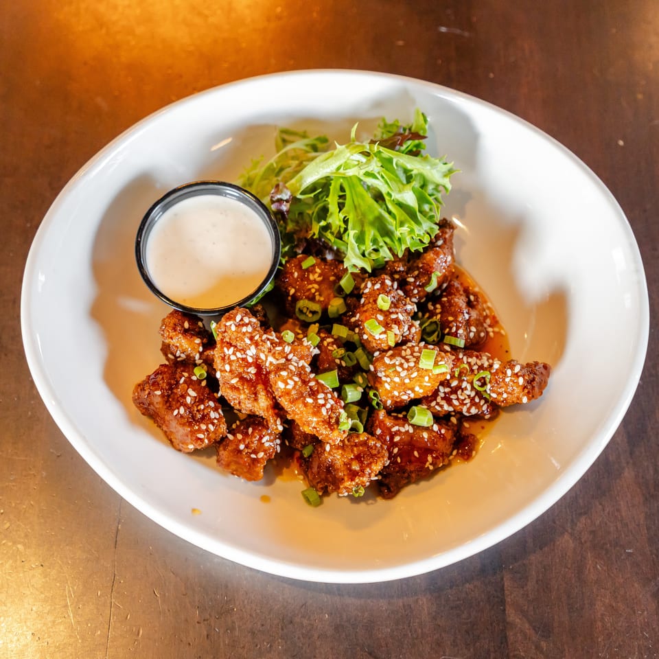 Glazed fried chicken pieces topped with sesame seeds and green onions, served with leafy greens and creamy dipping sauce.