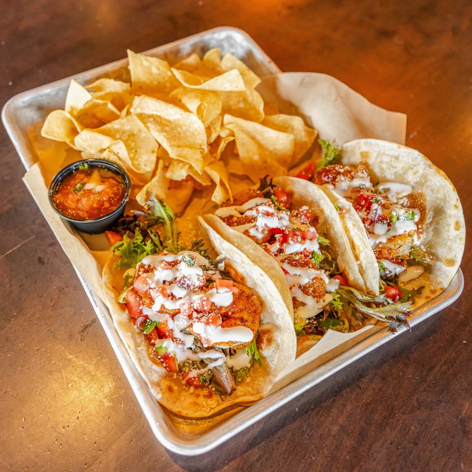 Tray of three loaded tacos with creamy sauce, salsa, greens, tortilla chips, and dipping salsa on a wooden table.
