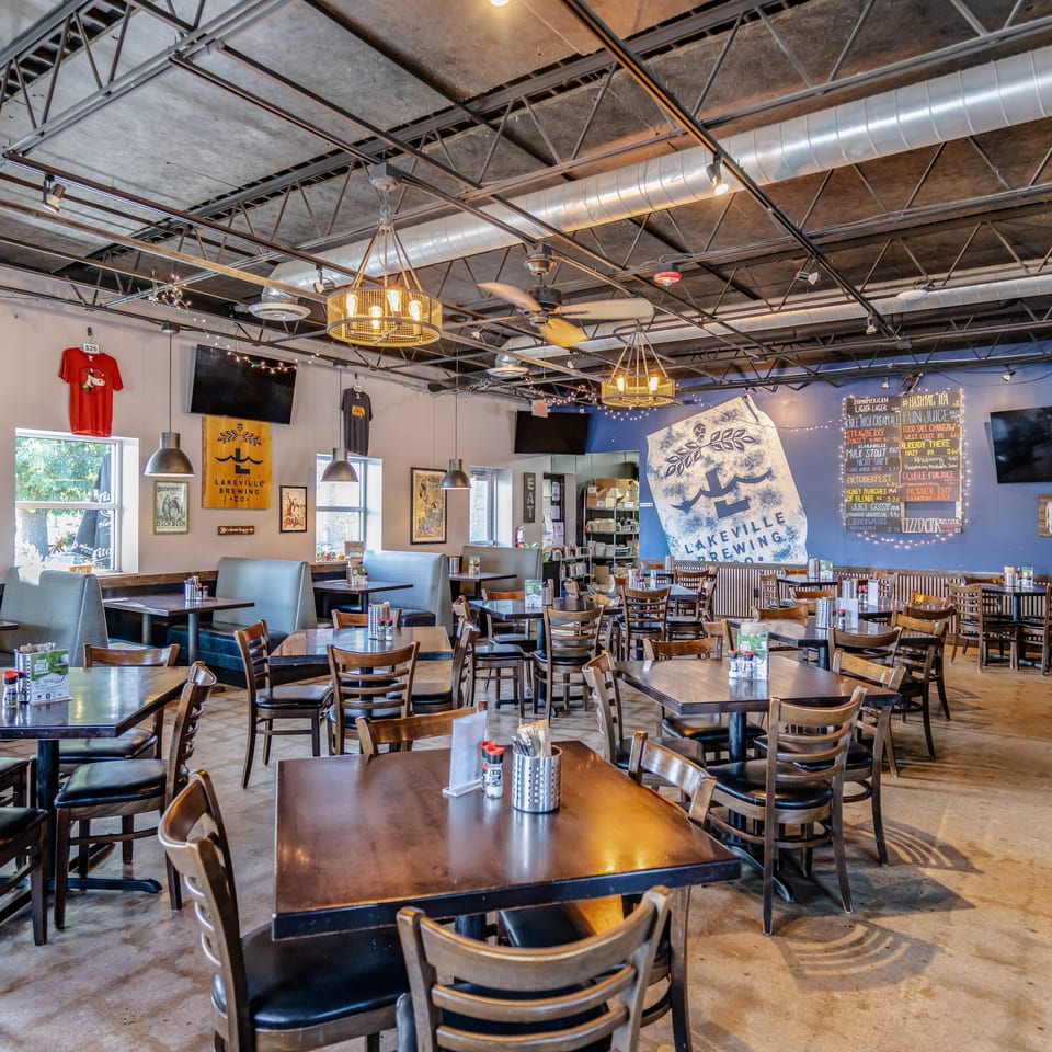 Spacious Lakeville Brewing Co. taproom with wooden tables, chairs, exposed beams, industrial ductwork, and a chalkboard beer menu.