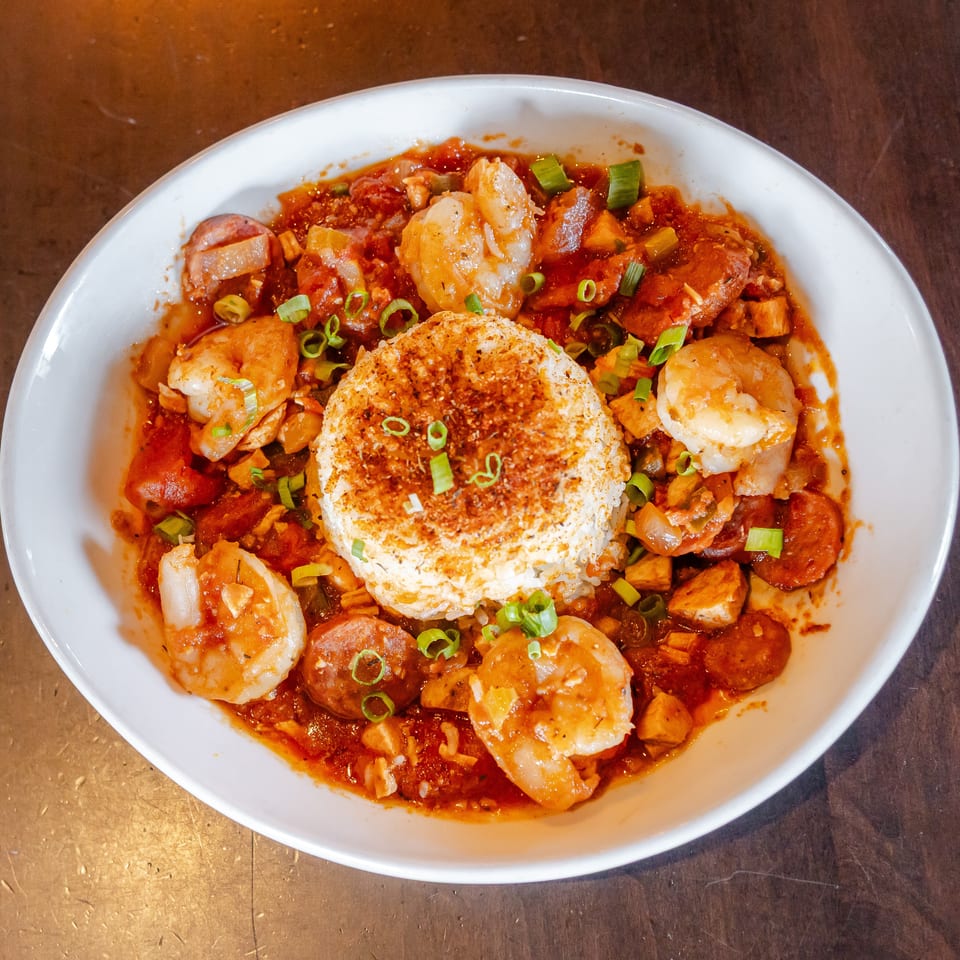 Bowl of shrimp, sausage, and vegetables in red sauce surrounding a seasoned mound of rice, garnished with green onions.