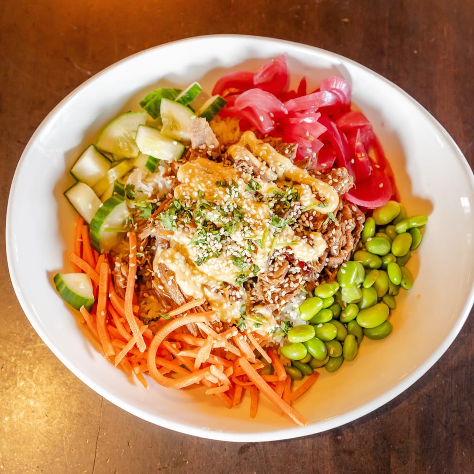 Colorful rice bowl topped with shredded meat, sesame seeds, creamy sauce, carrots, cucumber, edamame, tomatoes, and pickled onions.