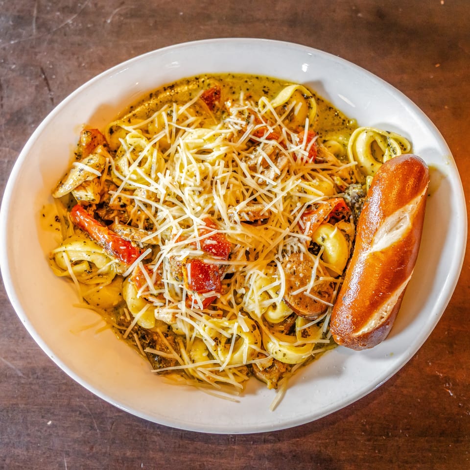 Creamy tortellini pasta with chicken, sun-dried tomatoes, shredded cheese, and bread in a white bowl.