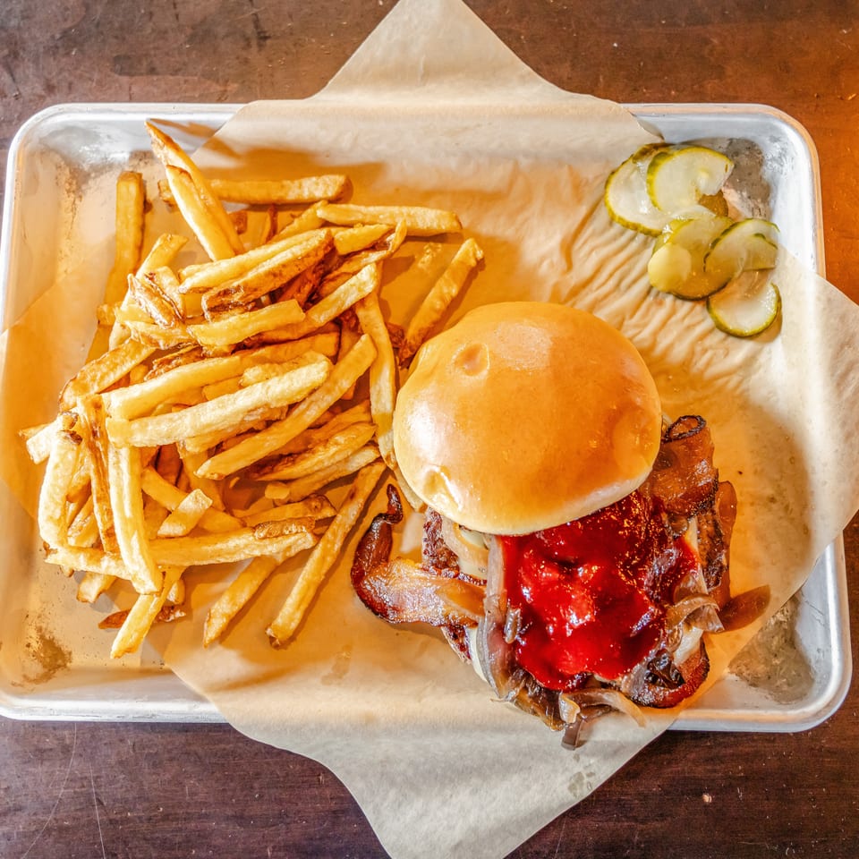 Bacon cheeseburger with barbecue sauce, onions, fries, and pickle slices served on a paper-lined metal tray.