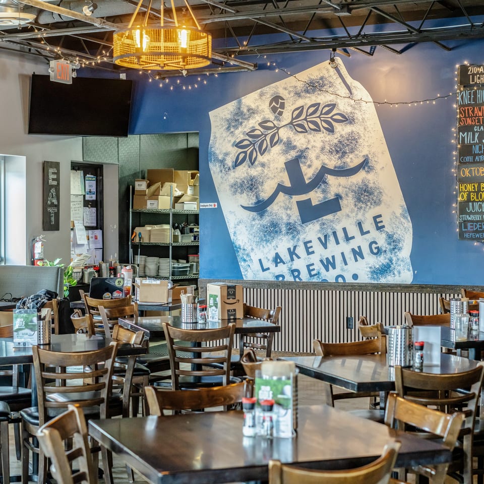 Lakeville Brewing restaurant interior with wooden tables, chairs, string lights, brewery mural, and chalkboard beer menu.