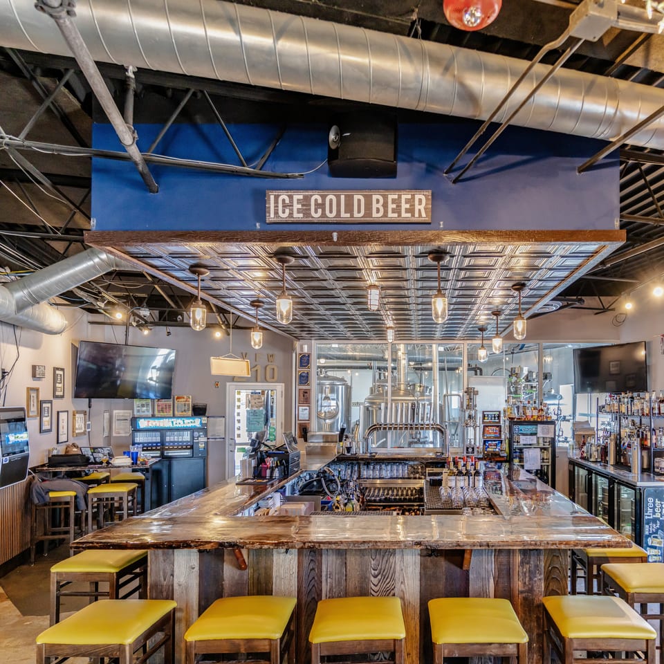 Industrial-style brewery bar with yellow stools, polished counter, brewing tanks, liquor shelves, and an illuminated “Ice Cold Beer” sign.