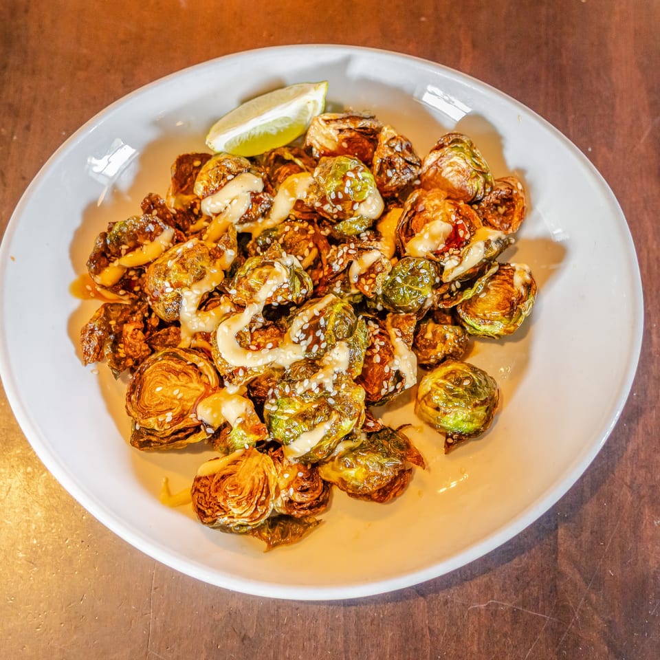 Roasted Brussels sprouts drizzled with creamy sauce and sprinkled with sesame seeds, served with a lime wedge on a white plate.